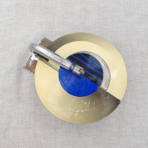 Peut inclure: Broche vintage au design circulaire. La broche présente un anneau extérieur crème, un centre bleu avec des rayures noires et bleues et un fermoir en métal argenté. Le design est géométrique et rétro.