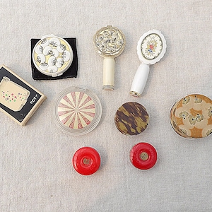 May include: A collection of vintage cosmetic compacts and accessories including a rectangular Coty Duette box, two handheld mirrors with floral and ornate designs, a circular red and white striped compact, and several round powder cases in various patterns.