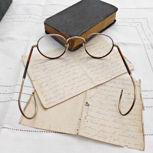 Puede incluir: Gafas redondas vintage con montura dorada y patillas marrones sobre cartas manuscritas antiguas. Un libro peque&ntilde;o y oscuro en el fondo. La escena evoca la historia y los estudios.