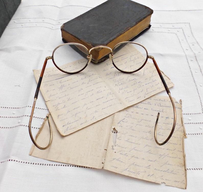 Puede incluir: Gafas redondas vintage con montura dorada y patillas marrones sobre cartas manuscritas antiguas. Un libro peque&ntilde;o y oscuro en el fondo. La escena evoca la historia y los estudios.