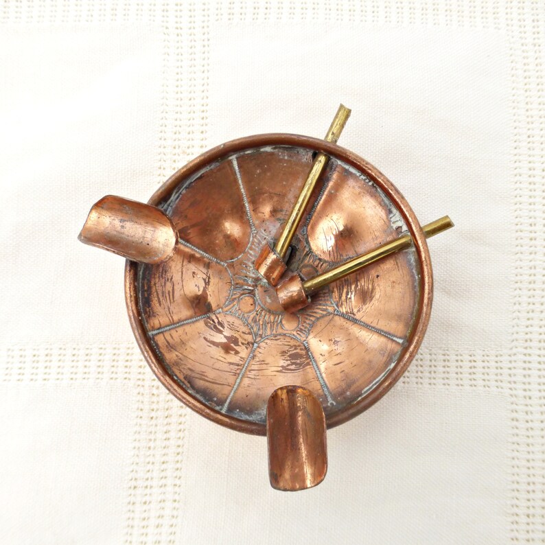 May include: A copper ashtray with three brass arms extending from the center. The arms are designed to hold cigarettes. The ashtray has a decorative design on the bottom.