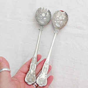 Pode incluir: Um par de utensílios de servir antigos em prata, um garfo e uma colher, com detalhes florais e de folhas ornamentados. As alças têm um design decorativo e curvo. Os utensílios são segurados em uma mão contra um fundo branco.