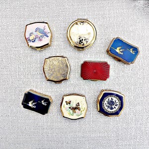 May include: A collection of eight small metal pill boxes with hinged lids. Designs include blue birds on a branch, a gold floral pattern, flying ducks on blue, a solid red sunburst, a butterfly on cream, and a blue and white floral medallion.