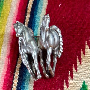 May include: A silver-toned metal brooch featuring two horses in a side-by-side running pose. The horses have detailed manes and tails with a smooth, metallic finish. The brooch is designed with a pin mechanism on the back to secure it to fabric.