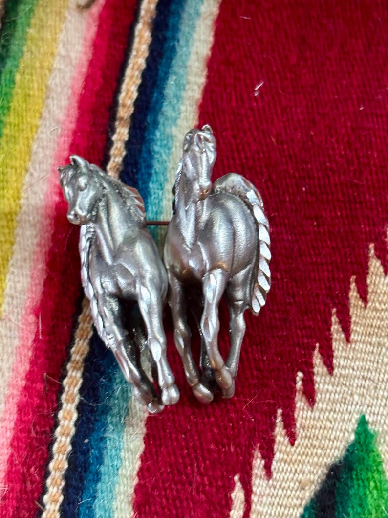 May include: A silver-toned metal brooch featuring two horses in a side-by-side running pose. The horses have detailed manes and tails with a smooth, metallic finish. The brooch is designed with a pin mechanism on the back to secure it to fabric.