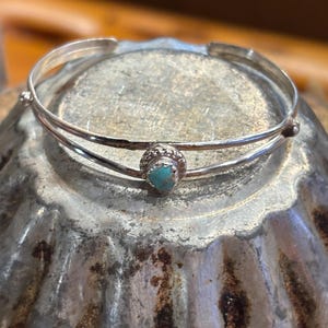 May include: A silver-toned cuff bracelet with a turquoise stone centerpiece. The bracelet has a double-band design with a textured finish and small decorative accents. The turquoise stone is round and set in a detailed bezel.