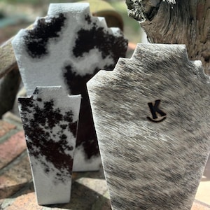 May include: Three cowhide necklace display stands. Two are white with black spots and one is gray with white streaks. The gray stand has a letter "K" logo.