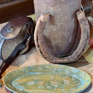 May include: An oval, green and blue ceramic platter with etched Southwestern designs. A rusty horseshoe and a leather strap are in the background, along with a weathered metal container. The items evoke a rustic, Western theme.