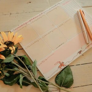May include: A weekly planner with a pink and beige design, featuring sections for each day and a decorative illustration. Two pens rest on the planner. Sunflowers with orange and yellow petals and green leaves are in the foreground.