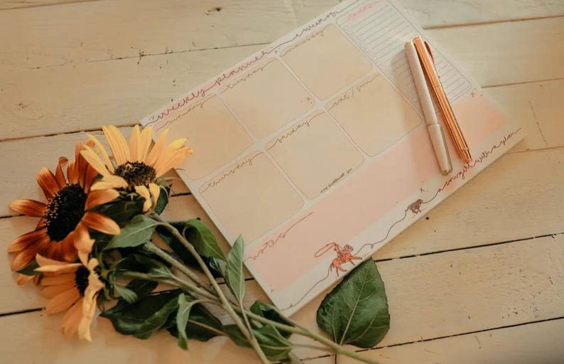 May include: A weekly planner with a pink and beige design, featuring sections for each day and a decorative illustration. Two pens rest on the planner. Sunflowers with orange and yellow petals and green leaves are in the foreground.