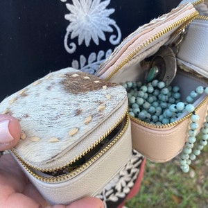 May include: Two small, square, zippered jewelry boxes. The top box is made of white cowhide with brown spots. The bottom box is a solid light pink. The bottom box is open and contains a turquoise necklace and a silver charm.
