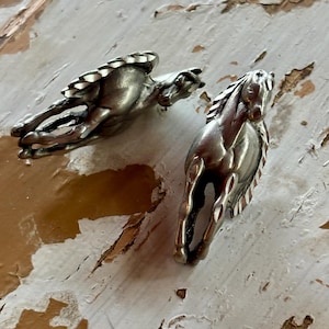 May include: Two metallic silver-toned horse figurines depicted in a dynamic running pose. Each horse features detailed manes and tails with a smooth, polished finish. The figures are sculpted with a sense of motion, showing the horses in mid-stride with their legs extended and heads held high, capturing a classic equine form.