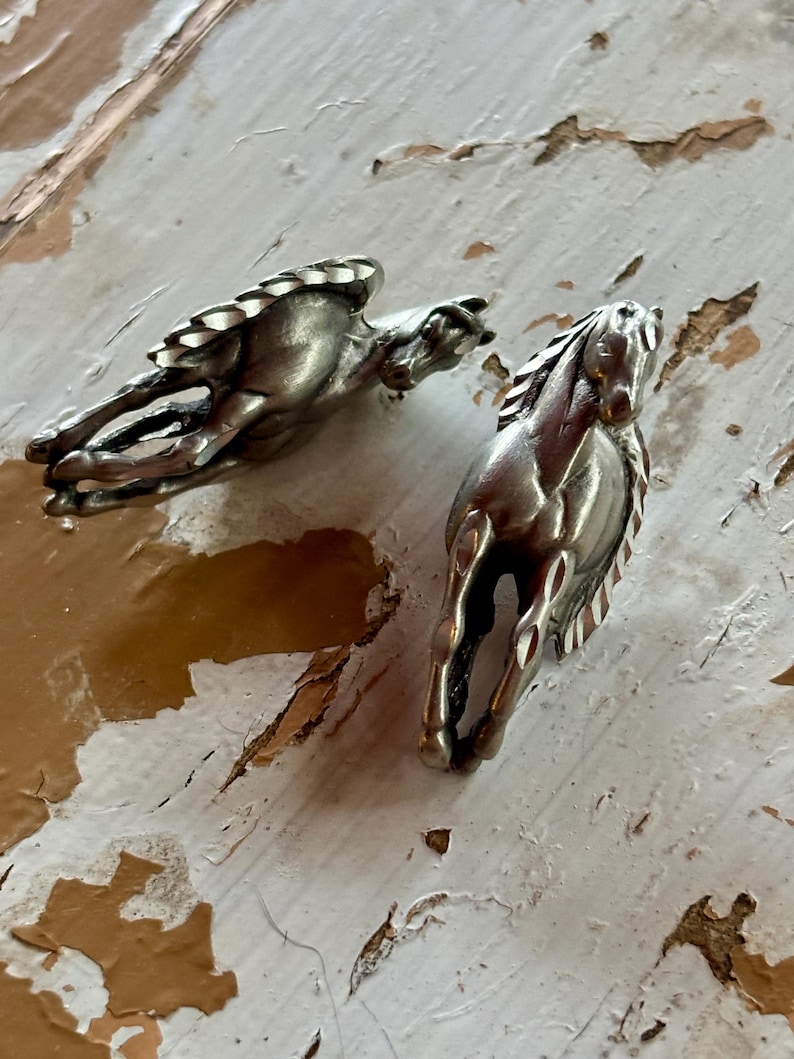 May include: Two metallic silver-toned horse figurines depicted in a dynamic running pose. Each horse features detailed manes and tails with a smooth, polished finish. The figures are sculpted with a sense of motion, showing the horses in mid-stride with their legs extended and heads held high, capturing a classic equine form.