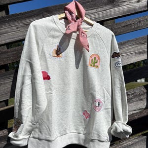 May include: A light gray sweatshirt with several colorful patches sewn on. The patches feature images of a cactus, a cowboy boot, a cowboy hat, and the word "Texas". A pink bandana is tied around the neck of the sweatshirt.