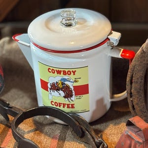 May include: White enamel coffee pot with a red trim and handle. The pot features a "Cowboy Coffee" label with a cowboy riding a horse. A clear glass knob sits atop the lid. Other rustic items are in the background.