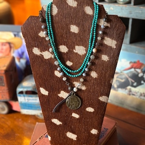 May include: A layered necklace displayed on a brown and white spotted stand. The top layer is turquoise beads, the middle layer has gray and white beads, and the bottom layer has a coin pendant with a leather cord.