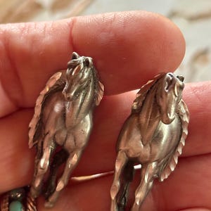 May include: A pair of silver-toned metal earrings shaped like running horses. Each earring features a detailed horse figure with a textured mane and tail, captured in a forward-facing galloping pose with a polished metallic finish.