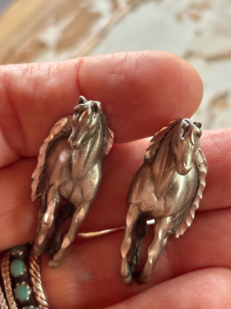 May include: A pair of silver-toned metal earrings shaped like running horses. Each earring features a detailed horse figure with a textured mane and tail, captured in a forward-facing galloping pose with a polished metallic finish.