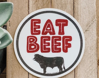 May include: White round sticker with a black border. The sticker says "EAT BEEF" in red letters with a black outline. A black silhouette of a bull is at the bottom of the sticker with the text "LEMON LORRAINE'S" underneath.