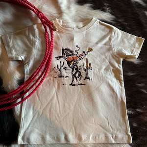 May include: A cream colored t-shirt with a graphic of a cowboy riding a bucking bronco in front of cacti.