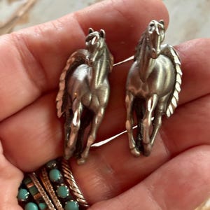 May include: A pair of silver-toned metal horse-shaped earrings featuring a detailed mane and running posture. The earrings have a polished finish and are presented alongside a multi-band ring featuring small, round, light blue turquoise stones.