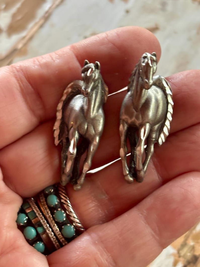 May include: A pair of silver-toned metal horse-shaped earrings featuring a detailed mane and running posture. The earrings have a polished finish and are presented alongside a multi-band ring featuring small, round, light blue turquoise stones.