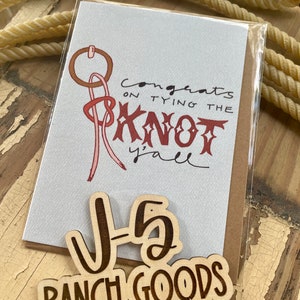 May include: A white card with the text "Congrats on tying the knot y'all" and a red and brown illustration of a lasso. Two wooden signs with the text "J-5 Ranch Goods" in brown lettering.