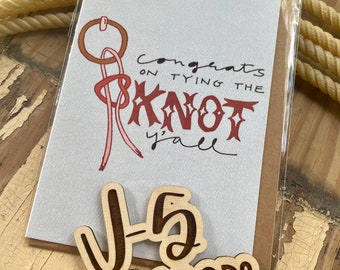 May include: A white card with the text "Congrats on tying the knot y'all" and a red and brown illustration of a lasso. Two wooden signs with the text "J-5 Ranch Goods" in brown lettering.