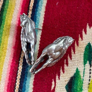 May include: Two silver-toned metal horse pins. One horse is depicted in a standing position, while the other horse is shown in a dynamic, running pose. Both figures feature detailed manes and muscular definition, crafted with a metallic finish.