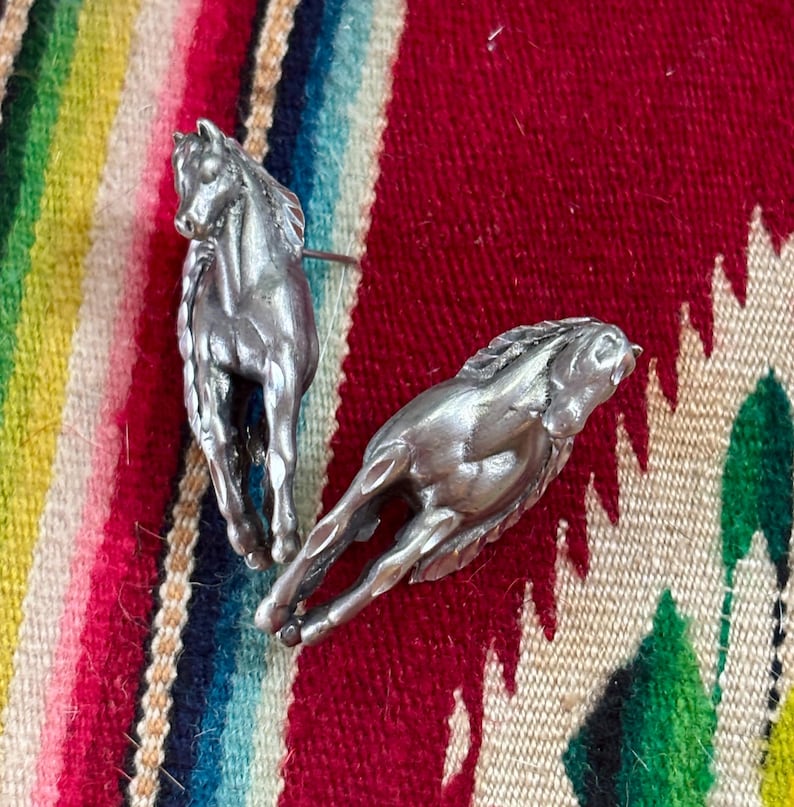 May include: Two silver-toned metal horse pins. One horse is depicted in a standing position, while the other horse is shown in a dynamic, running pose. Both figures feature detailed manes and muscular definition, crafted with a metallic finish.
