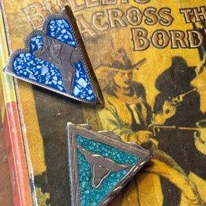 May include: Two silver-toned triangular pins with turquoise and blue inlay. Each pin features a bull silhouette. The pins rest on a vintage book with the title "Bullets Across the Border".