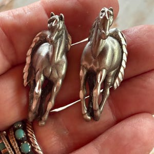 May include: A pair of silver-toned metal earrings shaped like running horses. Each horse figure features a textured, faceted mane and tail with a polished finish, capturing the animal in a forward-facing, galloping pose with detailed musculature.