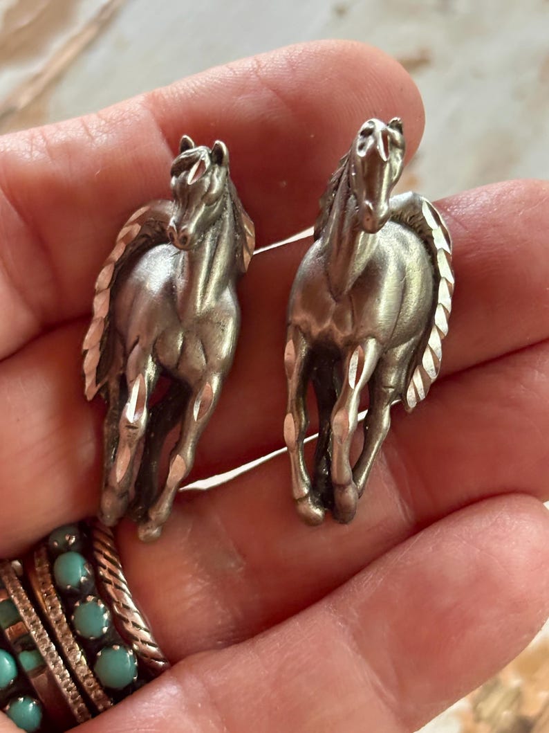 May include: A pair of silver-toned metal earrings shaped like running horses. Each horse figure features a textured, faceted mane and tail with a polished finish, capturing the animal in a forward-facing, galloping pose with detailed musculature.