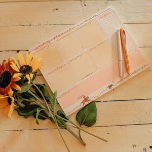 May include: A weekly planner with a peach and white design, featuring spaces for each day and a to-do list. Two pens and a bouquet of sunflowers rest on a light wood surface.