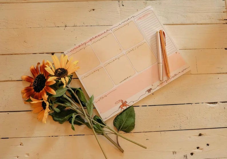 May include: A weekly planner with a peach and white design, featuring spaces for each day and a to-do list. Two pens and a bouquet of sunflowers rest on a light wood surface.