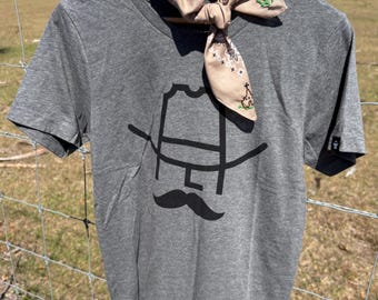May include: A heather grey t-shirt with a black graphic of a cowboy hat and mustache. The shirt is hanging on a wooden fence post with a brown bandana tied around the neck.