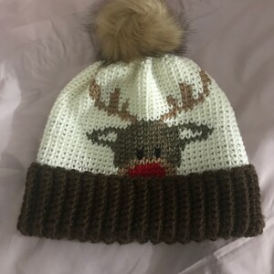 Op de afbeelding: Witte gebreide beanie met een bruin rendierontwerp en een bruine nep bont pompon bovenop.