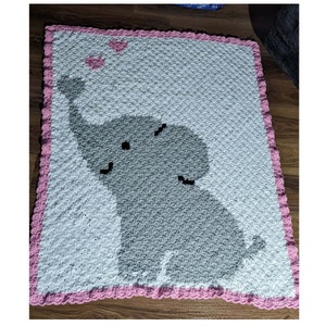 May include: A white and gray crocheted baby blanket with a pink border. The blanket features a gray elephant with pink hearts above it.