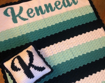 Könnte beinhalten: Eine weiße und schwarze gehäkelte Decke mit einem grün-weißen Streifenmuster. Die Decke trägt den Namen "Kennedi", der oben in Grün gehäkelt ist, und ein passendes Kissen mit dem Buchstaben "K", der in Schwarz gehäkelt ist.