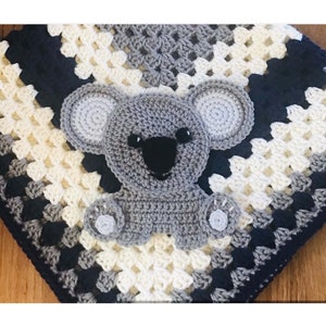 May include: A crocheted baby blanket with a gray koala applique. The blanket is made in a granny square pattern with alternating stripes of white, gray, and dark blue yarn.