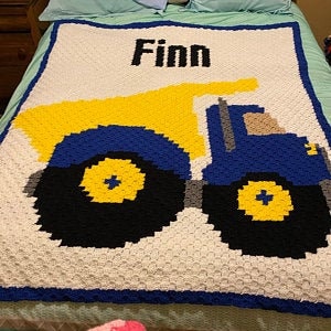 May include: A white crocheted baby blanket with a blue and yellow dump truck design. The blanket is personalized with the name "Finn".
