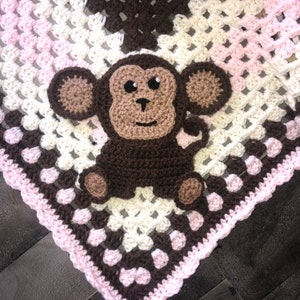 May include: A crocheted baby blanket with a brown monkey applique in the center. The blanket is made with white, pink, and brown yarn and has a scalloped edge.