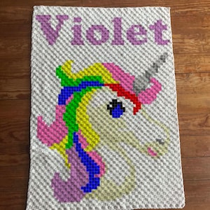 May include: A crocheted blanket with a unicorn design and the name "Violet" in purple. The unicorn has a rainbow mane, a white body, a pink horn, and blue eyes. The blanket is made of white yarn.