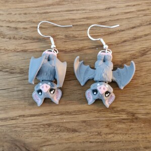 Könnte beinhalten: Ein Paar graue Fledermaus-Ohrringe mit silbernen Haken. Die Fledermäuse sind kopfüber, mit rosa Nasen und Ohren. Die Flügel sind ausgebreitet, und die Ohrringe sind vor einem Holzgrund platziert.