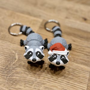 Könnte beinhalten: Zwei graue Waschbär-Schlüsselanhänger. Ein Waschbär trägt eine rote Weihnachtsmütze.