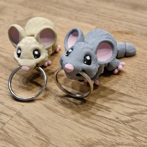 Könnte beinhalten: Zwei 3D-gedruckte Maus-Schlüsselanhänger, einer grau und einer beige, mit rosa Ohren und Nasen. Beide Schlüsselanhänger haben silberne Schlüsselringe.