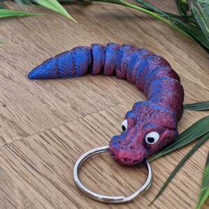 Könnte beinhalten: Ein lila und blauer Alligator-Schlüsselanhänger mit einem silbernen Schlüsselring. Der Alligator ist zu einem Kreis zusammengerollt.