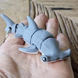 Könnte beinhalten: Ein grauer, 3D-gedruckter Hammerhai-Schlüsselanhänger. Der Hai hat einen segmentierten Körper, Flossen und einen Schlüsselring. Die Augen des Hais sind schwarz und weiß. Der Schlüsselanhänger wird in einer Hand gehalten.