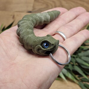 Könnte beinhalten: Ein grüner, 3D-gedruckter, raupenförmiger Schlüsselanhänger mit einem blauen Auge und einem silbernen Schlüsselring.