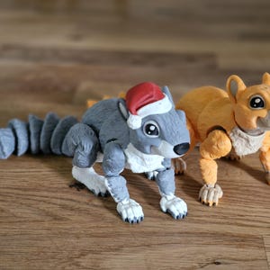 Könnte beinhalten: Zwei bewegliche Eichhörnchenfiguren. Eines ist grau mit rotem Weihnachtsmannhut, das andere ist orange. Beide haben detaillierte Pfoten und Schwänze. Sie sind auf einer Holzoberfläche platziert.